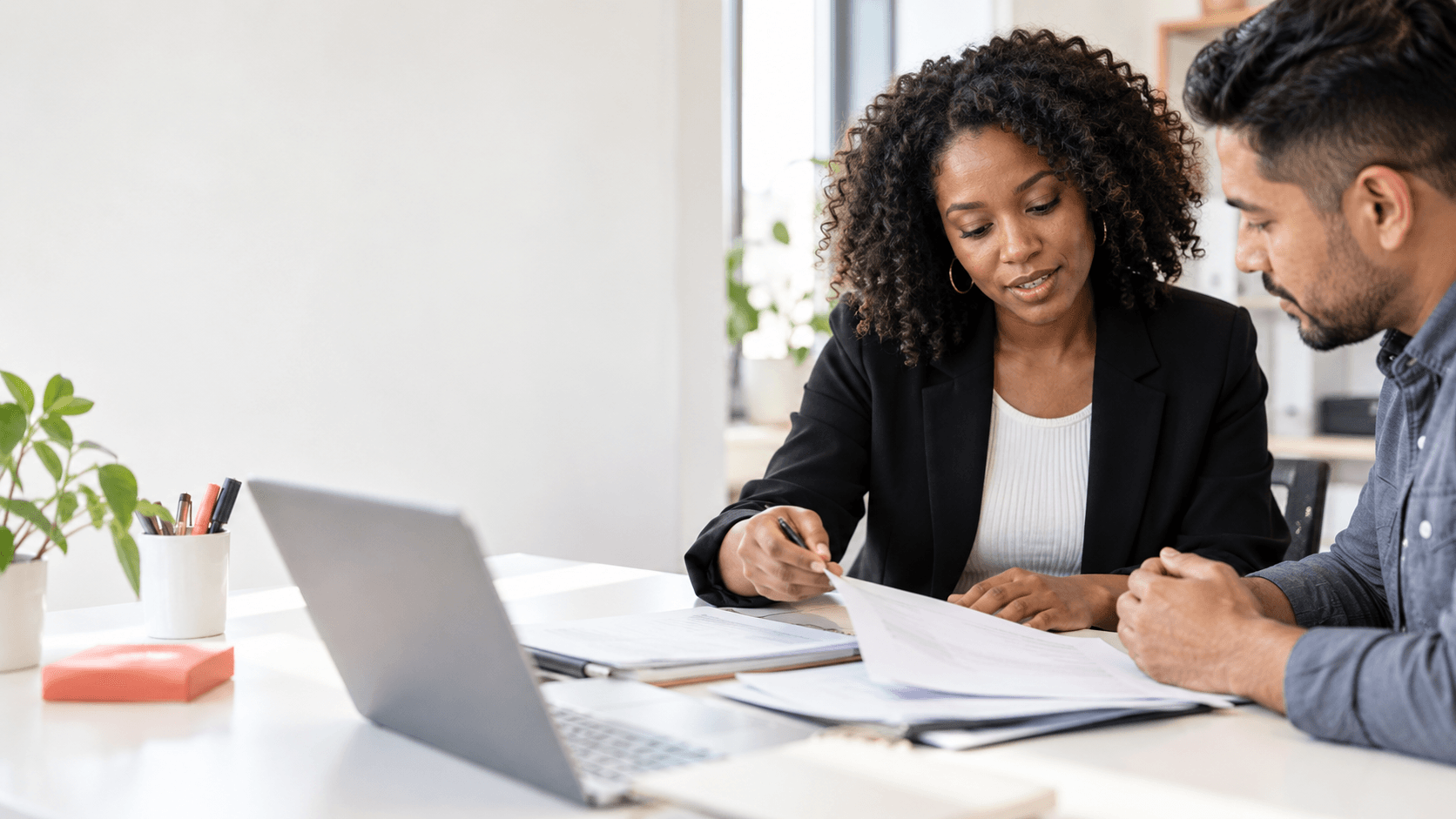Professional woman in black blazer reviewing documents with man at white desk with laptop